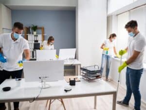 Team using effective cleaning techniques to sanitize office desks and computer equipment while wearing masks and gloves.