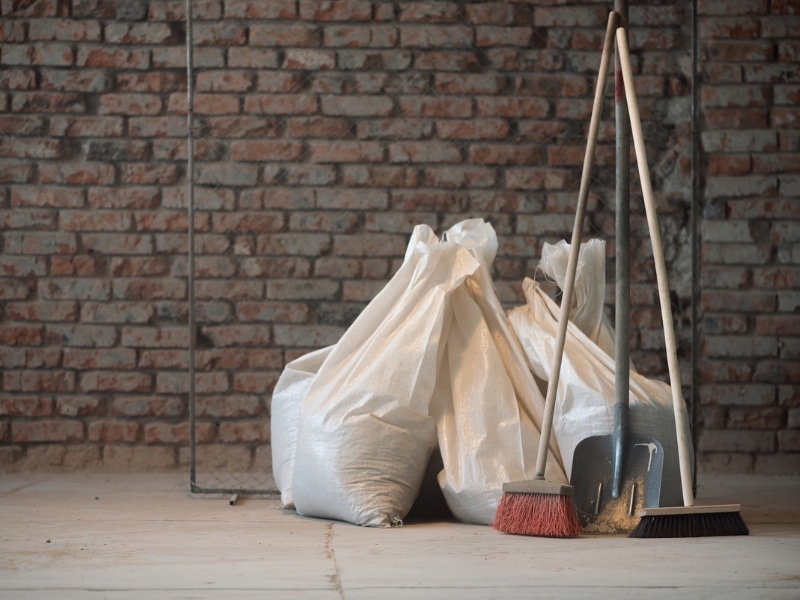 Clean up construction site tools and debris bags stacked against exposed brick wall during post-construction cleaning.